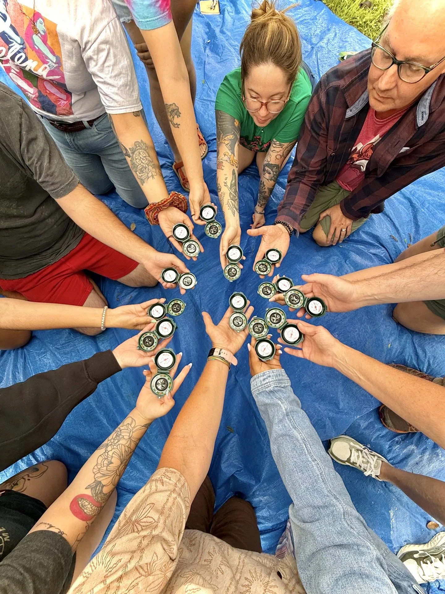 A group seated in a circle outdoors, hands together over a blue tarp.